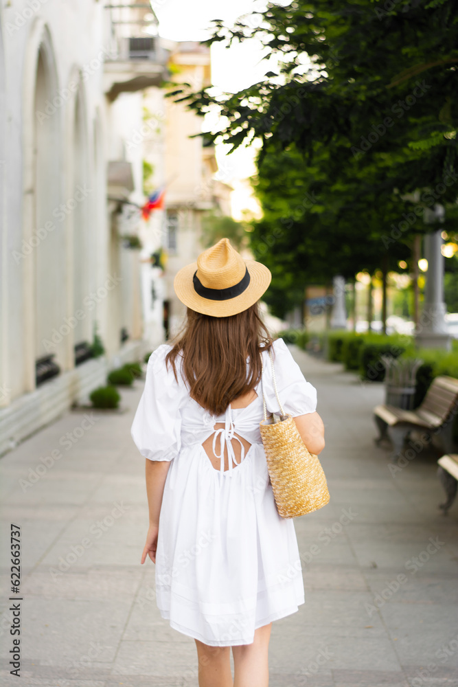 Back view a tourist woman in a white fashionable trendy cotton dress short with a straw hat, a straw bag, walks in a European city. Architecture, vacation, stylish dress, fashion, 20-30 years