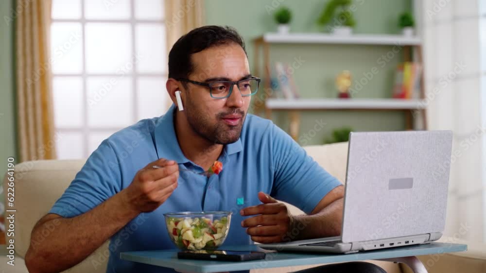 Happy indian man eating fruits salad while talking on online video call at home - concept of healthy eating, Video conference and Remote conversation