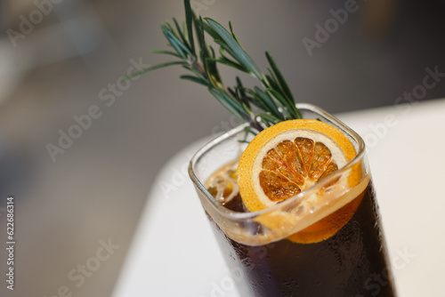 a glass of ice coffee with dried orange and rosemary, on white table in cafe or restaurant