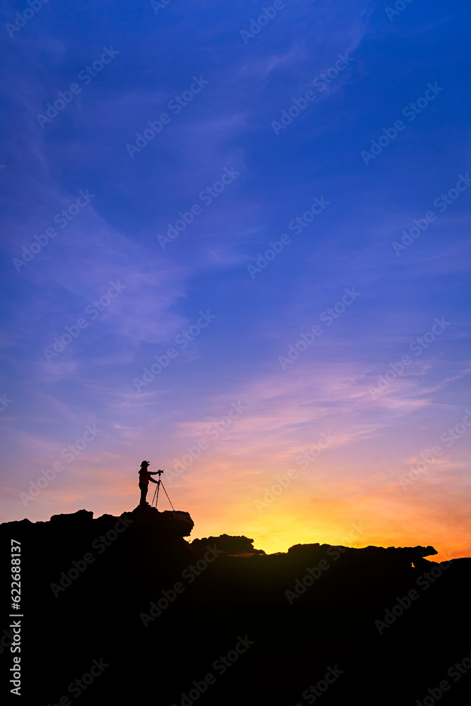 Silhouette of a man photographer standing on top of mountain at sunrise background. The photographer raised hands with the tripod takes photos high in the mountains.