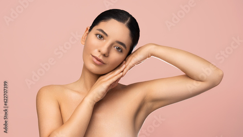 Beauty portrait of multiethnic attractive woman with serene expression holding palms to her face on pink background