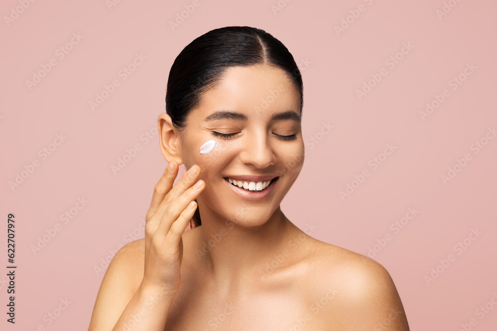 Beauty portrait. Attractive multi-ethnic woman 25s with cheerful smile applying cream on face for skincare