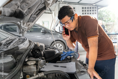 An Asian male car mechanic using his phone while working on repairing a car's engine at car repair garage