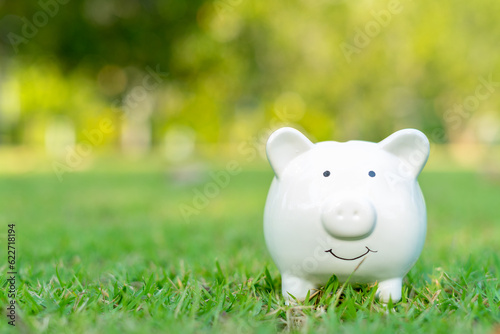 Investing and saving money concept. Cute white piggy bank standing in green grass outdoors with copy space. Financial planning and money saving concept.