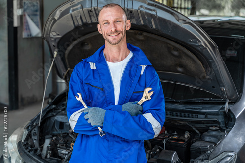 A happy male car mechanic in blue jumpsuit holding a wranch in each hands and cross his arms while standing in front of a car with hood open photo