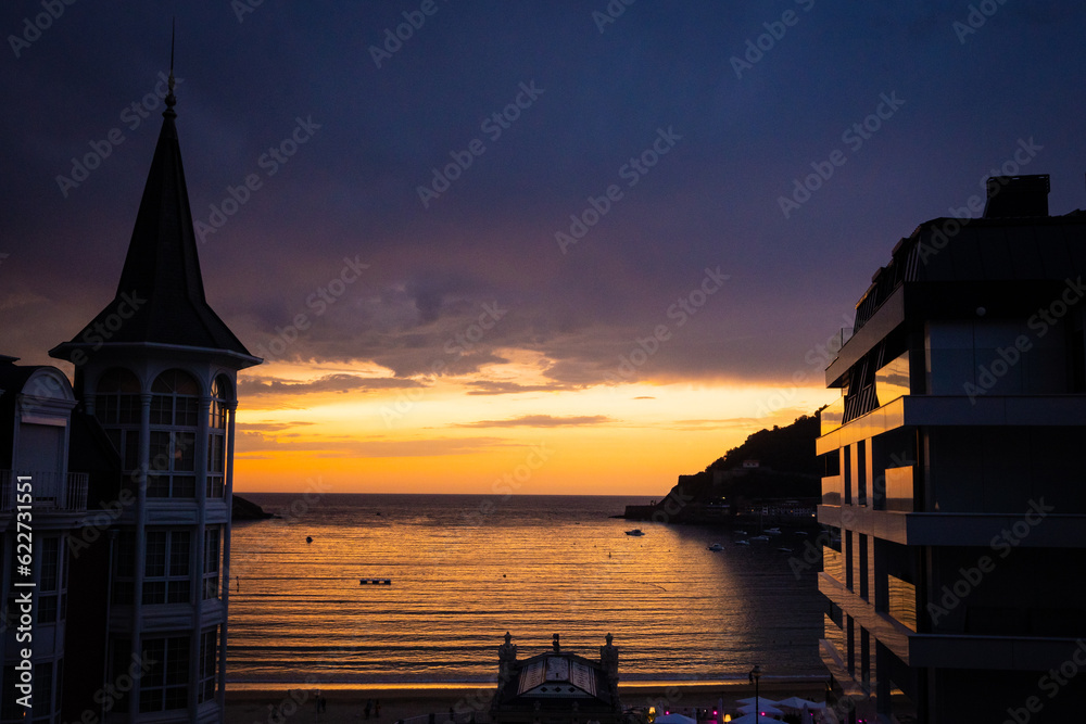 Naklejka premium Evening sunset view of bahia de la concha Donosita-San Sebastian Spain at dusk twilight. romantic colours fill the sky and reflect off the water with boats moored at sea in the natural harbour bay