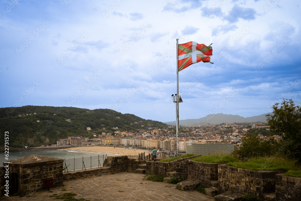 official flag of the Basque Country in Spain Ikurriña flies from Mount Urgull above Donosita San Sebastian on sunny summer day