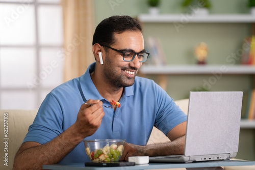 Happy indian man eating fruits salad while talking on online video call at home - concept of healthy eating  Video conference and Remote conversation