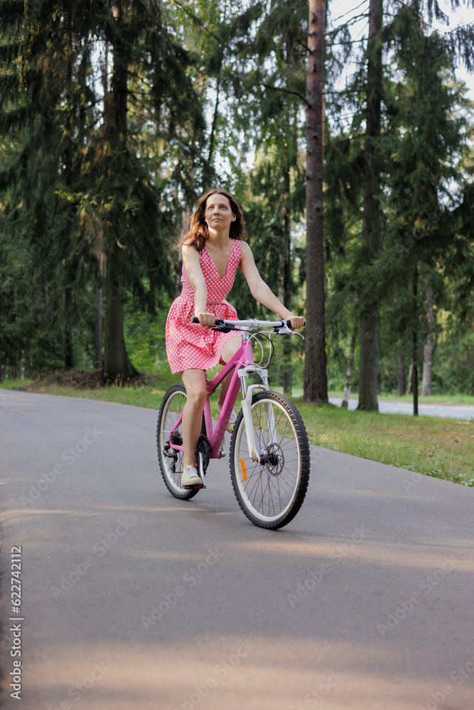 young active woman in pink dress rides a bicycle in park among the trees. Women fun weekend sports and recreation. Walking along bike path in national park, freedom and speed. riding