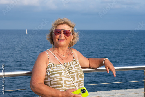 On a sunny day near the beach, an attractive elderly lady with white hair smiled as she read messages on her smart phone. The senior woman enjoyed her free time and vacation while holding the cellphon #622750919