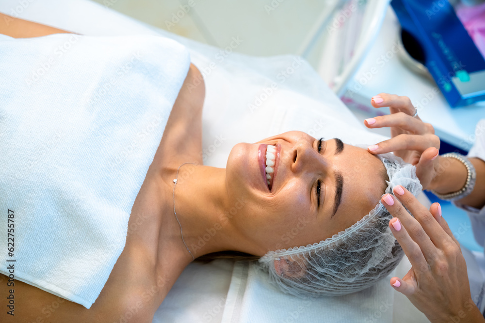 Fototapeta premium Cosmetologist preparing client for cosmetic procedures by putting disposable cap on woman head