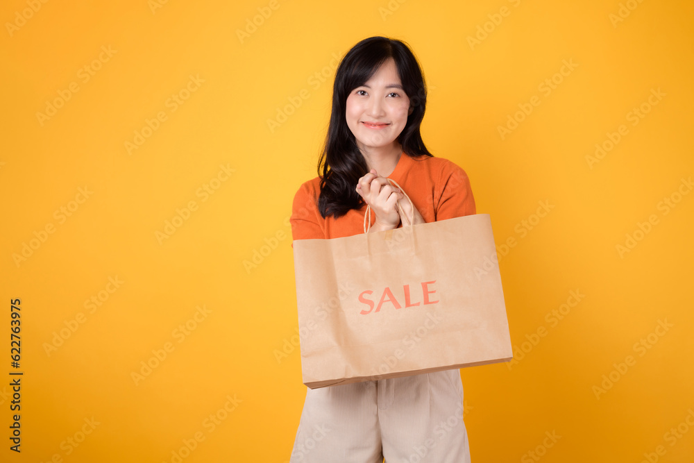 Portrait happy attractive 30s asian young woman with trendy springtime dress, orange shirt fashion and paper bag isolated on yellow background. Summertime sale shopping concept. Portrait happy attractive 30s asian young woman with trendy springtime dress, orange shirt fashion and paper bag isolated on yellow background. Summertime sale shopping concept.