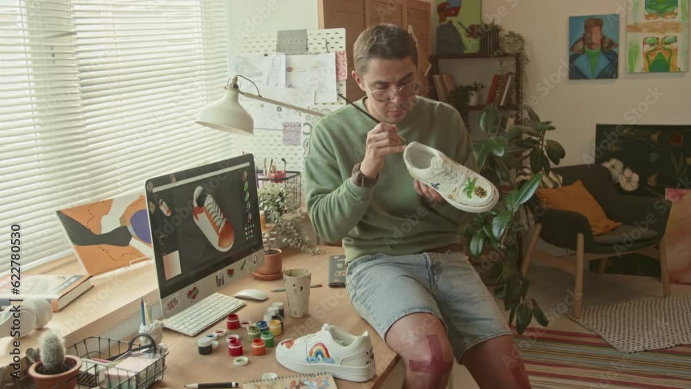 Medium full shot of male Caucasian designer sitting on corner of table in loft studio holding customized sneakers with colorful pictures and drawing on them at daytime
