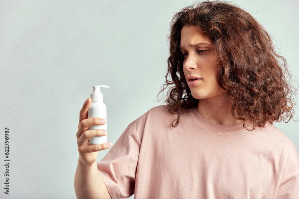 Young handsome long haired guy holding a bottle of facial wash or beauty cream in his hand. Caucasian man with brown hair uses hydrating skin products in his skincare routine to keep healthy look. Young handsome long haired guy holding a bottle of facial wash or beauty cream in his hand. Caucasian man with brown hair uses hydrating skin products in his skincare routine to keep healthy look.