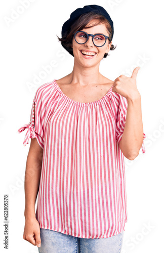 Beautiful young woman with short hair wearing casual clothes and glasses smiling with happy face looking and pointing to the side with thumb up.