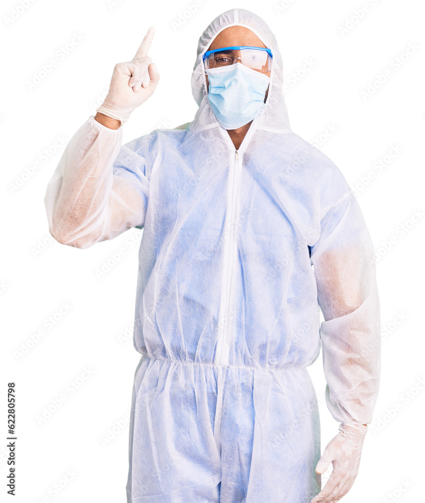 Young hispanic man wearing doctor protection coronavirus uniform and medical mask pointing finger up with successful idea. exited and happy. number one. Young hispanic man wearing doctor protection coronavirus uniform and medical mask pointing finger up with successful idea. exited and happy. number one.