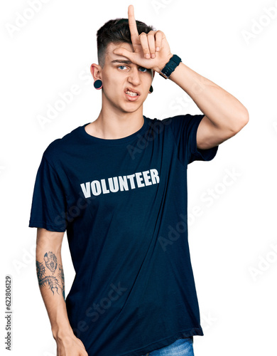 Young caucasian boy with ears dilation wearing volunteer t shirt making fun of people with fingers on forehead doing loser gesture mocking and insulting.