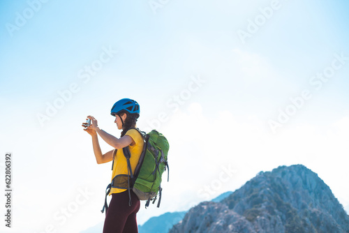 climber on top of the mountain takes a photo. Girl with a backpack and hard hat in the mountains. Woman traveler takes a photo on a smartphone, takes pictures of beautiful mountain scenery.