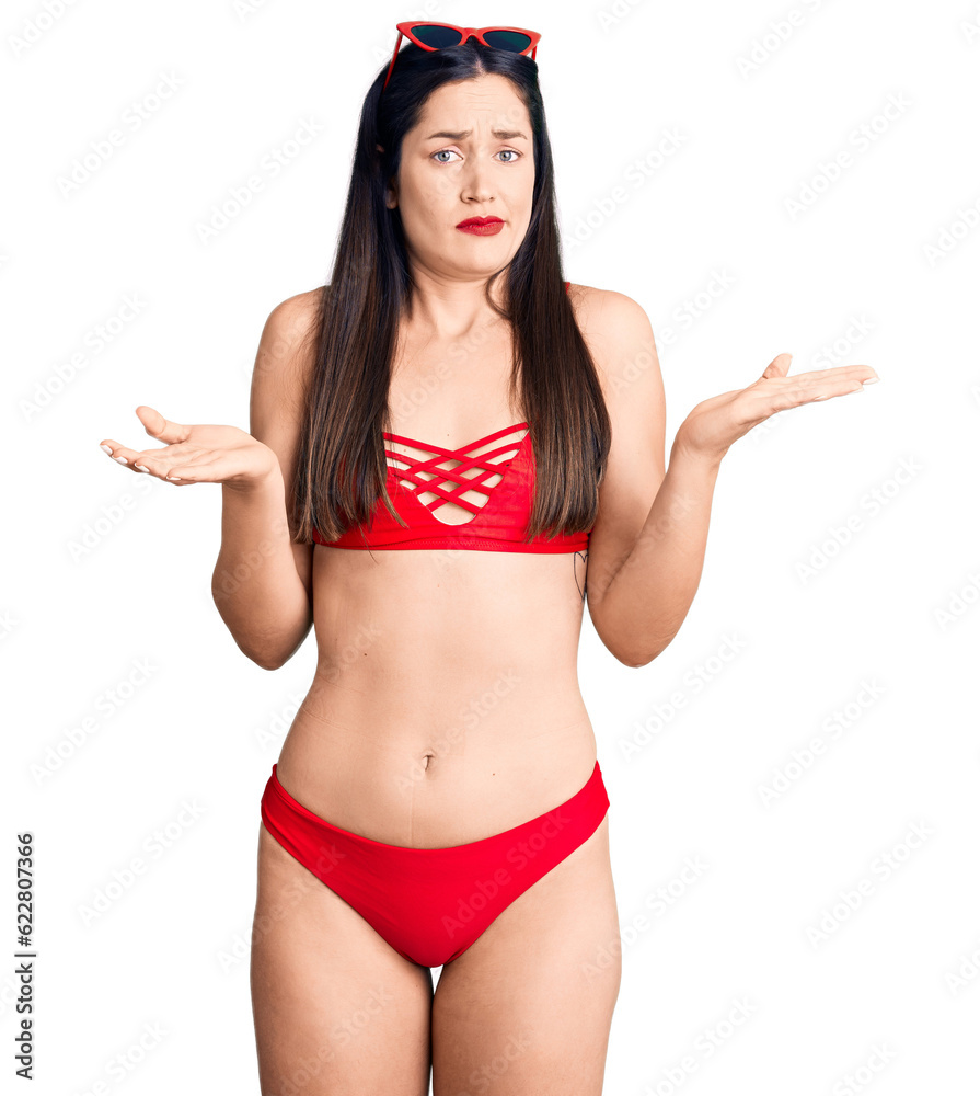 Young beautiful caucasian woman wearing bikini clueless and confused expression with arms and hands raised. doubt concept. Young beautiful caucasian woman wearing bikini clueless and confused expression with arms and hands raised. doubt concept.