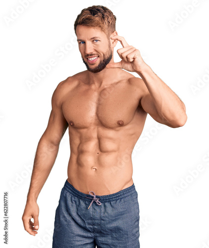 Young caucasian man standing shirtless smiling and confident gesturing with hand doing small size sign with fingers looking and the camera. measure concept.