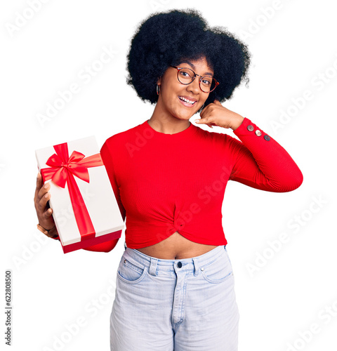 Young african american girl holding gift smiling doing phone gesture with hand and fingers like talking on the telephone. communicating concepts.