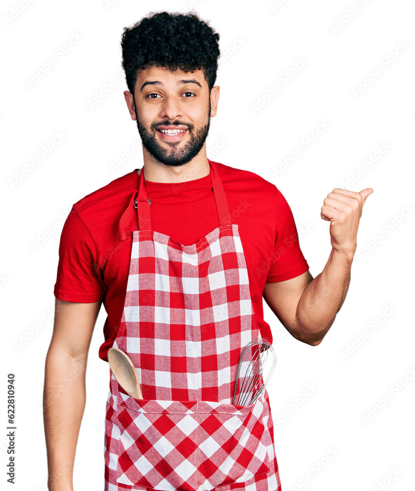Young arab man with beard wearing cook apron smiling with happy face looking and pointing to the side with thumb up. Young arab man with beard wearing cook apron smiling with happy face looking and pointing to the side with thumb up.
