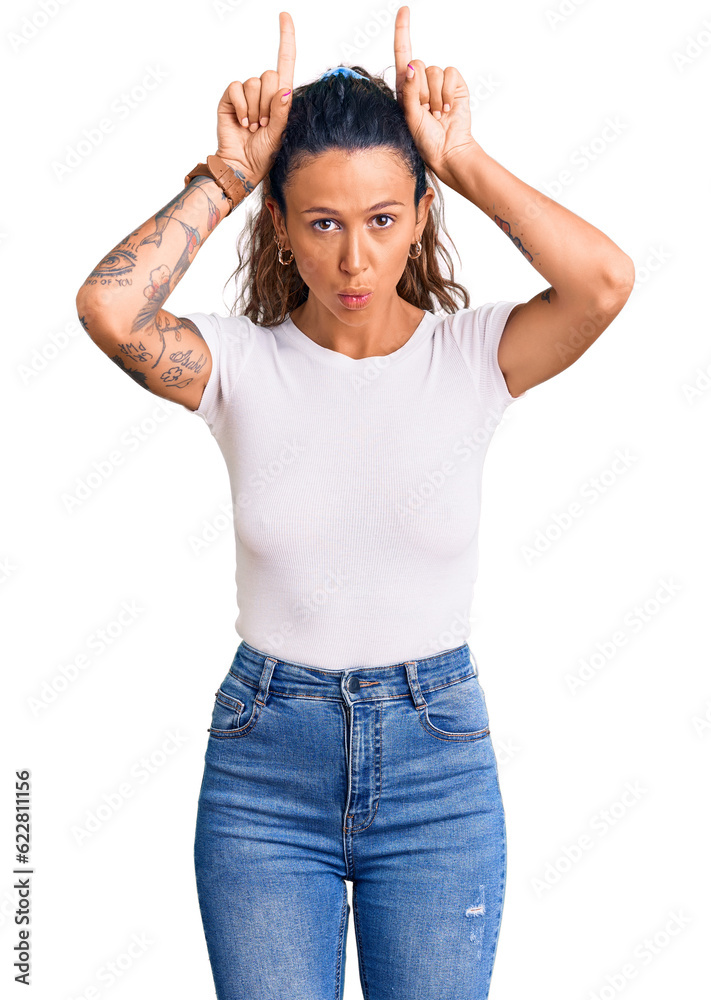 Young hispanic woman with tattoo wearing casual white tshirt doing funny gesture with finger over head as bull horns Young hispanic woman with tattoo wearing casual white tshirt doing funny gesture with finger over head as bull horns