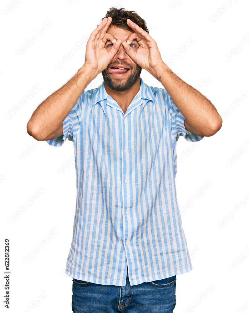 Handsome young man with beard wearing casual fresh shirt doing ok gesture like binoculars sticking tongue out, eyes looking through fingers. crazy expression. Handsome young man with beard wearing casual fresh shirt doing ok gesture like binoculars sticking tongue out, eyes looking through fingers. crazy expression.