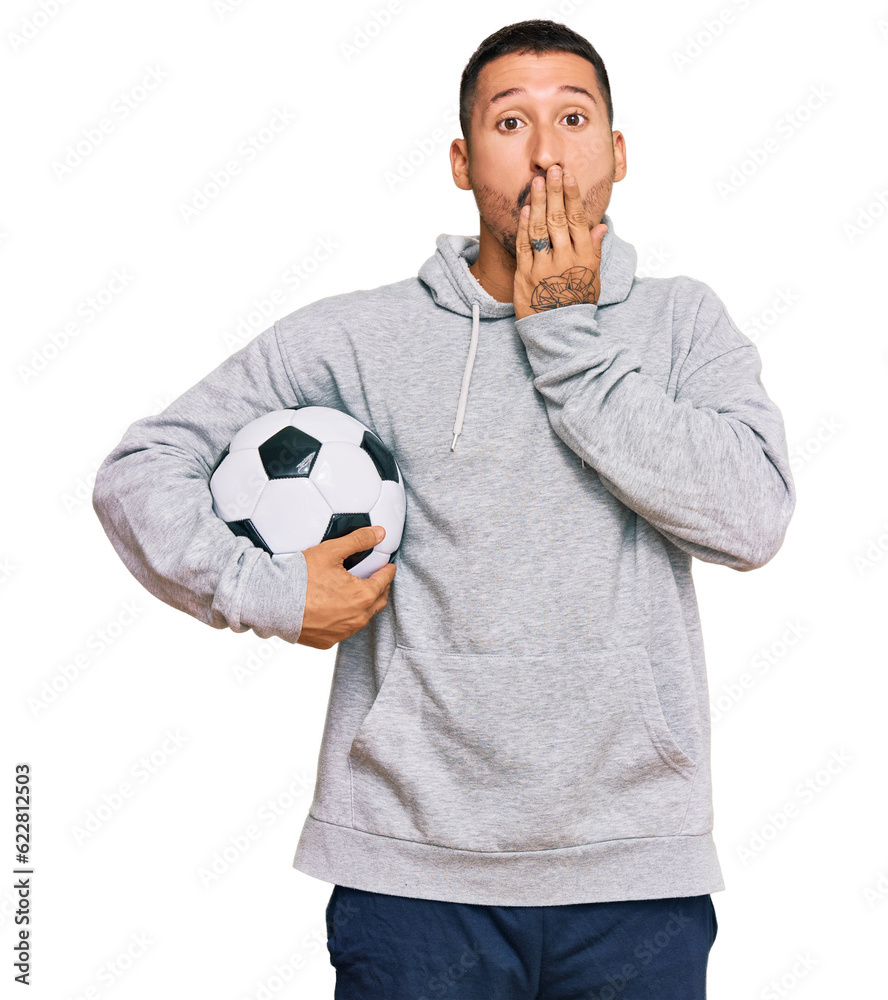 Handsome man with tattoos holding soccer ball covering mouth with hand, shocked and afraid for mistake. surprised expression Handsome man with tattoos holding soccer ball covering mouth with hand, shocked and afraid for mistake. surprised expression