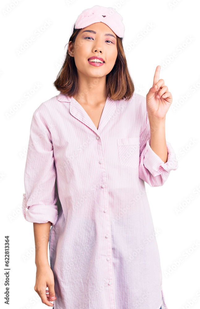 Young beautiful chinese girl wearing sleep mask and pajama showing and pointing up with finger number one while smiling confident and happy.