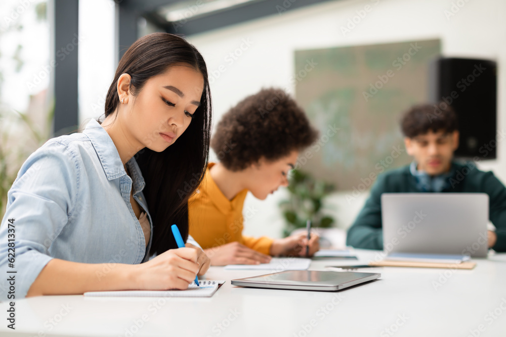 Asian lady sitting at desk and taking notes, writing essay, studying for class assignment with classmates on background