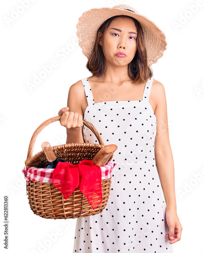 Fotografie Young beautiful chinese girl wearing summer hat holding picnic basket thinking a
