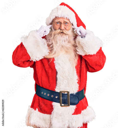 Old senior man with grey hair and long beard wearing traditional santa claus costume covering ears with fingers with annoyed expression for the noise of loud music. deaf concept.