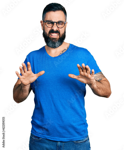 Hispanic man with beard wearing casual t shirt and glasses disgusted expression, displeased and fearful doing disgust face because aversion reaction. with hands raised