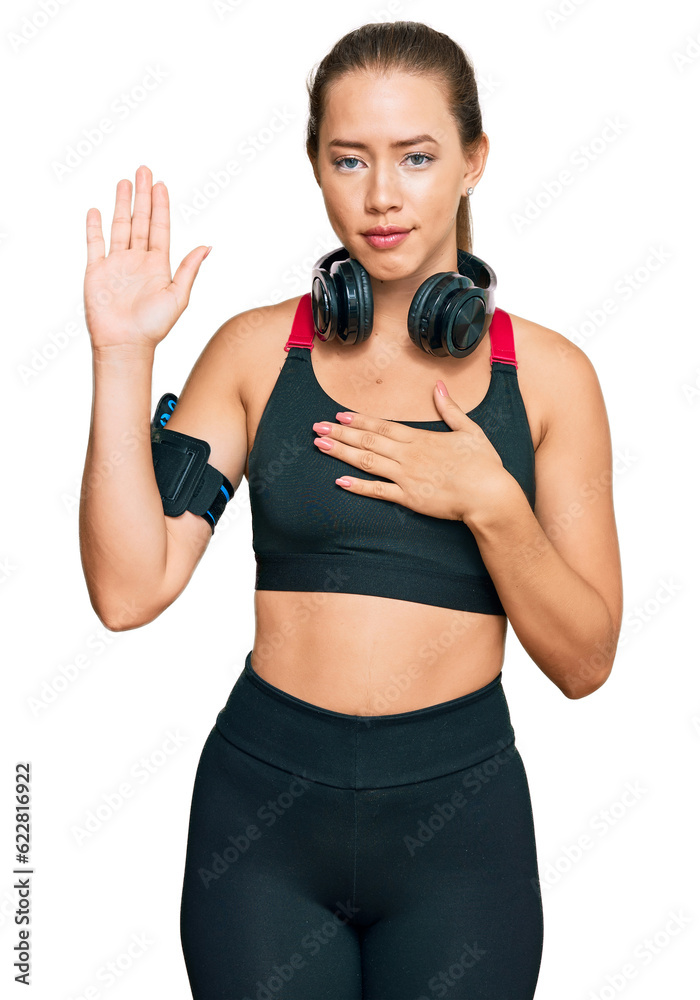 Beautiful blonde woman wearing gym clothes and using headphones swearing with hand on chest and open palm, making a loyalty promise oath Beautiful blonde woman wearing gym clothes and using headphones swearing with hand on chest and open palm, making a loyalty promise oath