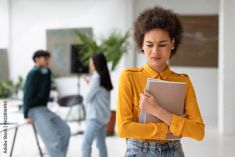 Portrait of unhappy and sad black female student with copybooks in hands, being bullying by her groupmates Portrait of unhappy and sad black female student with copybooks in hands, being bullying by her groupmates