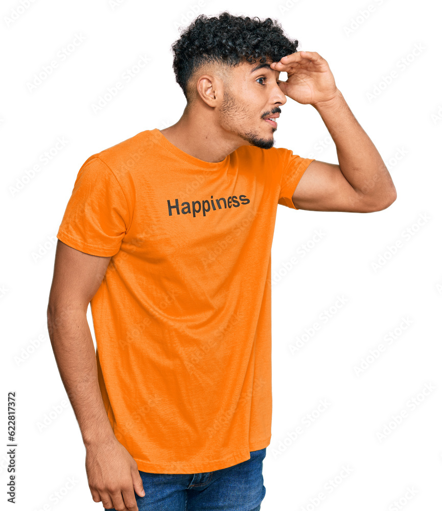 Young arab man wearing tshirt with happiness word message very happy and smiling looking far away with hand over head. searching concept. Young arab man wearing tshirt with happiness word message very happy and smiling looking far away with hand over head. searching concept.