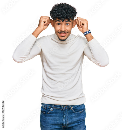 Young arab man wearing casual winter sweater smiling pulling ears with fingers, funny gesture. audition problem