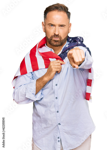 Handsome middle age man holding united states flag pointing with finger to the camera and to you, confident gesture looking serious