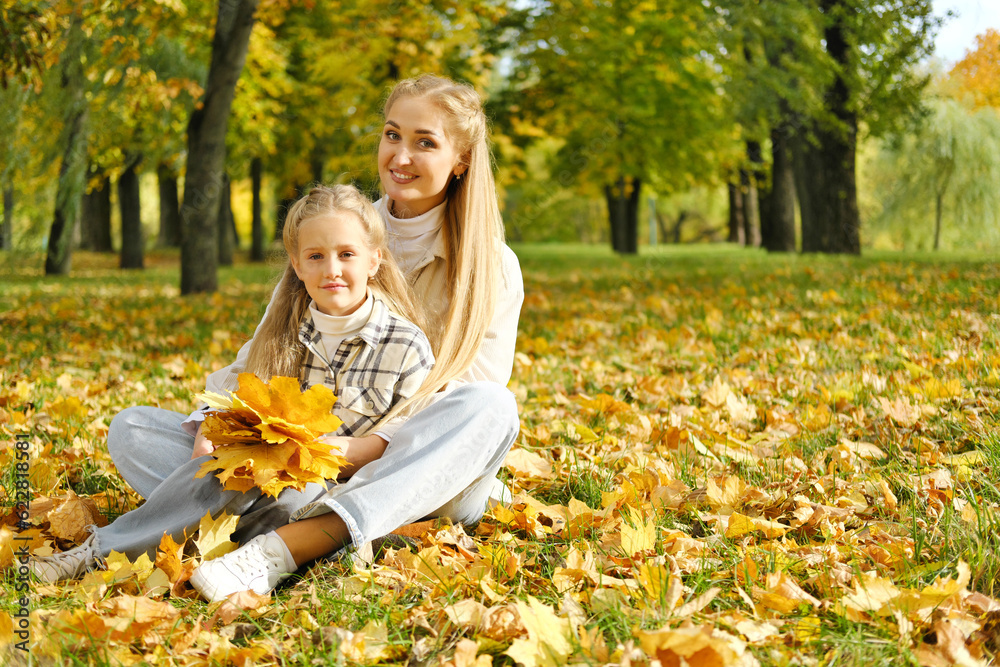 Fototapeta premium The family has fun in the park. Girl and mother sit on the ground among yellow leaves, smile and look at the camera