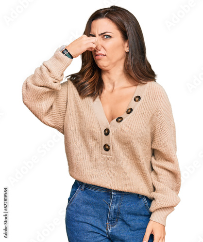 Young brunette woman wearing casual winter sweater smelling something stinky and disgusting, intolerable smell, holding breath with fingers on nose. bad smell