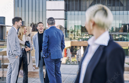 group of businessmen laughing and chatting while a woman in the foreground, out of focus, looks at them.