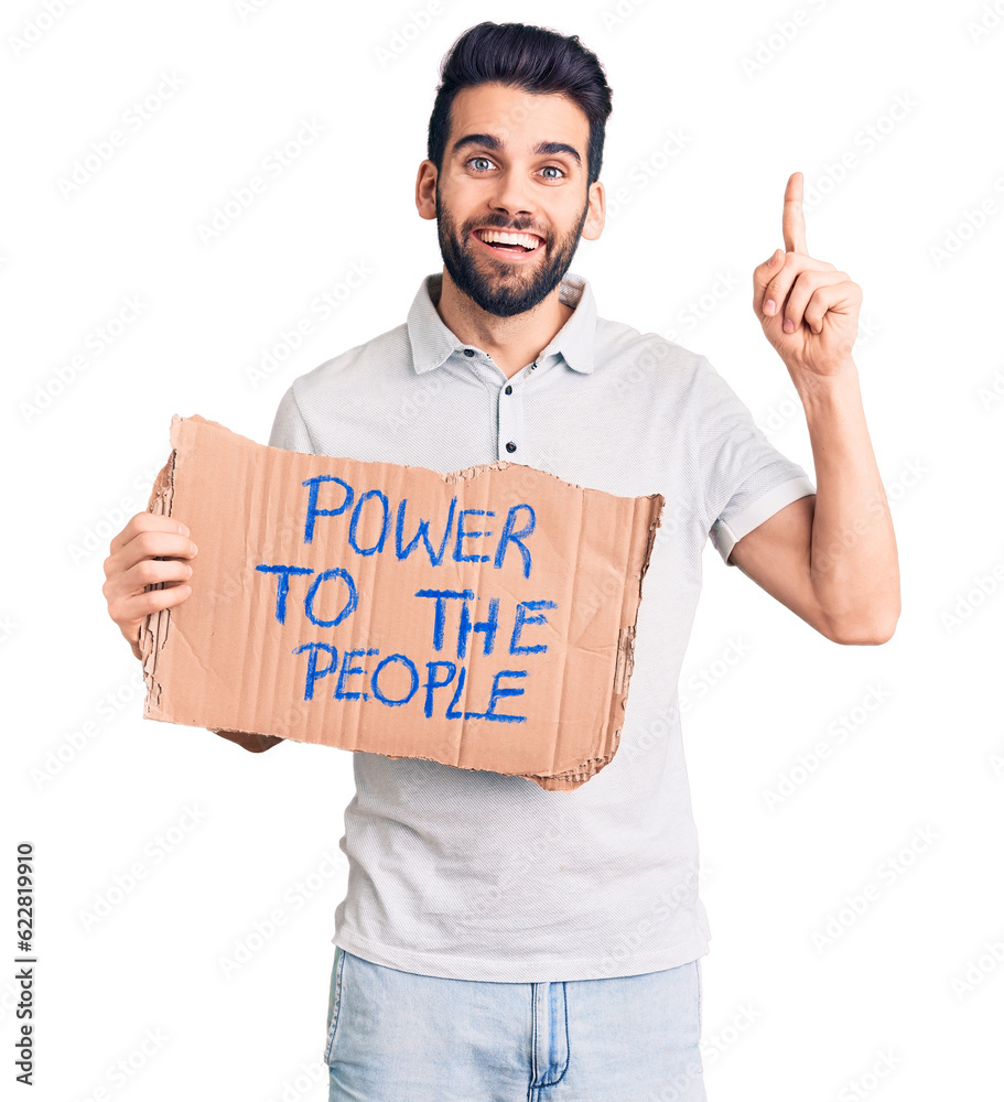 Young handsome man with beard holding power to the people cardboard banner surprised with an idea or question pointing finger with happy face, number one Young handsome man with beard holding power to the people cardboard banner surprised with an idea or question pointing finger with happy face, number one