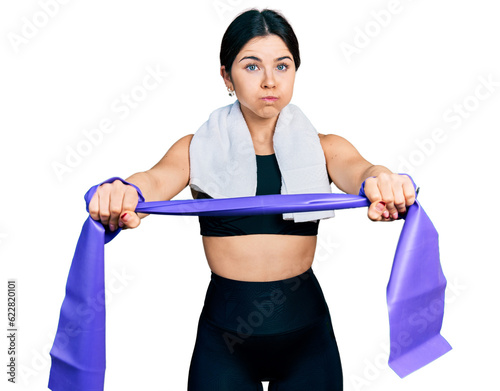 Young brunette woman with blue eyes training arm resistance with elastic arm bands puffing cheeks with funny face. mouth inflated with air, catching air.