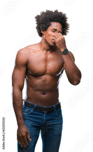 Afro american shirtless man showing nude body over isolated background smelling something stinky and disgusting, intolerable smell, holding breath with fingers on nose. Bad smells concept.