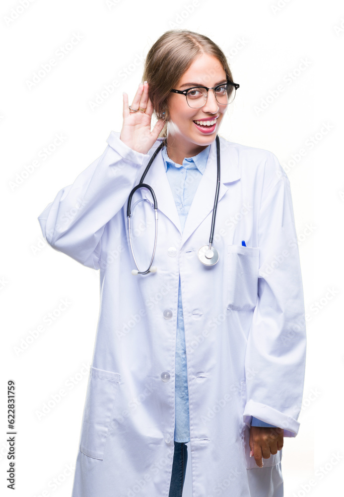 Beautiful young blonde doctor woman wearing medical uniform over isolated background smiling with hand over ear listening an hearing to rumor or gossip. Deafness concept.