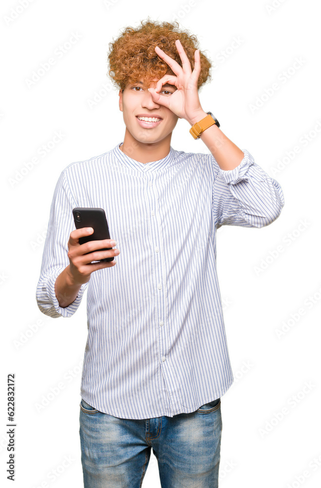 Young handsome man using smartphone with happy face smiling doing ok sign with hand on eye looking through fingers Young handsome man using smartphone with happy face smiling doing ok sign with hand on eye looking through fingers