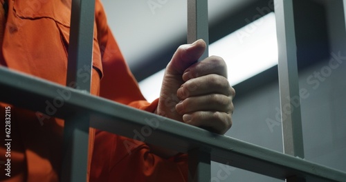 Close up of prisoner in orange uniform holding metal bars, standing in prison cell. Guilty criminal or killer serves imprisonment term for crime. Inmate in jail or detention center. Justice system.