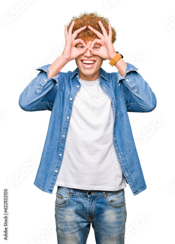 Young handsome man with afro hair wearing denim jacket doing ok gesture like binoculars sticking tongue out, eyes looking through fingers. Crazy expression.