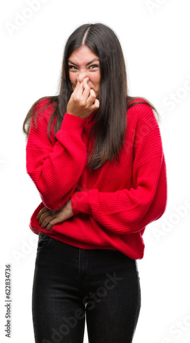 Young beautiful hispanic wearing red sweater smelling something stinky and disgusting, intolerable smell, holding breath with fingers on nose. Bad smells concept.